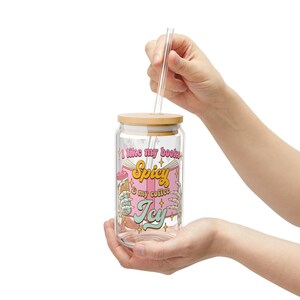 Pu&ograve; includere: Un barattolo di vetro trasparente con un coperchio in bamb&ugrave; e una cannuccia. Il barattolo ha un design rosa e bianco con il testo "Mi piacciono i miei libri Spicy & il mio caff&egrave; Icy".