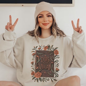 May include: A beige sweatshirt with a floral design and the text "Dibs on the Villain" printed on the front.