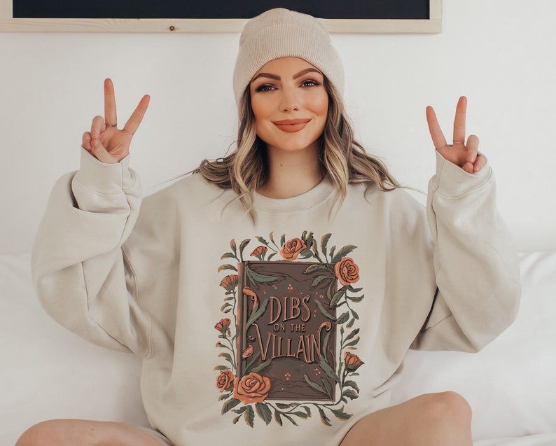May include: A beige sweatshirt with a floral design and the text "Dibs on the Villain" printed on the front.