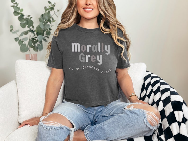 May include: A woman wearing a dark gray cropped t-shirt with the text "Morally Grey is my favorite color" printed on it. She is sitting on a white couch with a black and white checkered throw blanket.