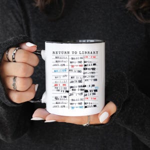 Puede incluir: Taza de cer&aacute;mica blanca con interior y asa negros, con el texto "RETURN TO LIBRARY" y una lista de fechas. La taza est&aacute; sujeta por una persona. Las fechas est&aacute;n impresas en una fuente de m&aacute;quina de escribir.