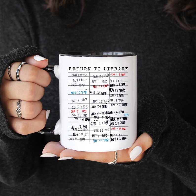 Puede incluir: Taza de cer&aacute;mica blanca con interior y asa negros, con el texto "RETURN TO LIBRARY" y una lista de fechas. La taza est&aacute; sujeta por una persona. Las fechas est&aacute;n impresas en una fuente de m&aacute;quina de escribir.
