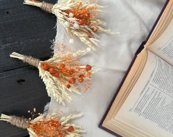 Puede incluir: Tres pequeños ramos rústicos de trigo seco, bayas naranjas y acentos blancos están dispuestos sobre una tela clara. Cada ramo está atado con hilo. Un libro abierto con páginas envejecidas está en el fondo, lo que sugiere un tema otoñal natural.