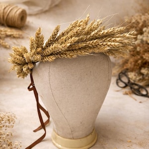 Peut inclure: Une couronne de blé, composée d'épis de blé séchés, est présentée sur une tête de mannequin. La couronne est fixée avec un ruban marron. La tête est en tissu beige et repose sur une base crème. Le fond comprend des fleurs séchées et une bobine de ficelle.