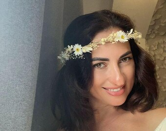 May include: A woman wearing a floral crown made of white daisies and dried flowers. She is smiling and wearing a cream-colored off-the-shoulder top. The background includes a gray curtain and a patterned wall.