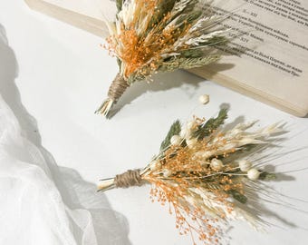 Puede incluir: Dos pequeños ramos de flores secas con acentos de trigo, naranja y verde, atados con hilo. Los ramos están dispuestos sobre una superficie blanca cerca de un libro abierto con texto.