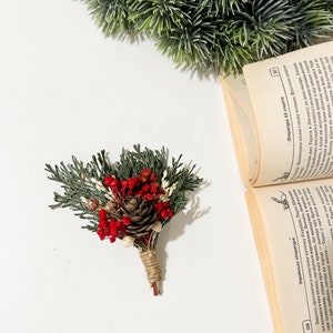 May include: A small bouquet of dried flowers with red berries, a pine cone, and green sprigs tied with twine. The bouquet is on a white surface.