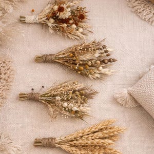 May include: Four small dried flower bouquets in a variety of textures and colours. The bouquets are tied with twine and lace, creating a rustic, natural look. The flowers include wheat, seed pods, and other dried elements, ideal for home decoration.