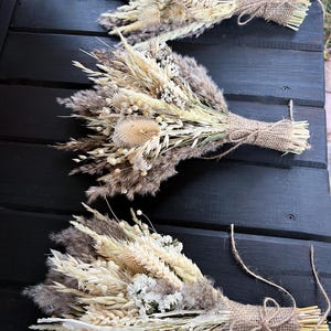 Può includere: Tre bouquet di fiori secchi con un'estetica rustica e naturale. Ogni bouquet è legato con spago e presenta un mix di erbe secche, grano e fiori bianchi.