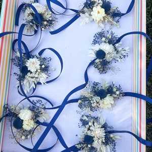 May include: Six blue ribbon wrist corsages with white dried flowers and blue accents in a striped box.