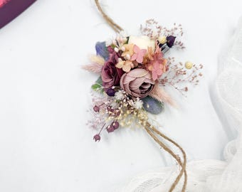 May include: A floral wrist corsage featuring a cluster of pink, purple, and cream-colored flowers, accented with greenery and small white blossoms. The arrangement is tied with a natural twine cord, perfect for weddings or special events.