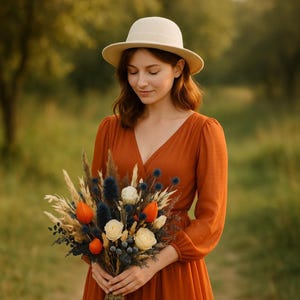Könnte beinhalten: Eine Frau mit cremefarbenem Hut und rostfarbenem Kleid hält einen Strauß Trockenblumen. Der Strauß enthält orange, weiße und blaue Blumen sowie getrocknete Gräser. Der Hintergrund ist ein verschwommenes grünes Feld.