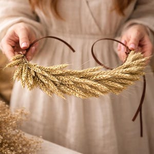 Könnte beinhalten: Ein handgefertigter Weizenkranz mit braunem Band, gehalten von einer Person. Der Kranz besteht aus getrockneten Weizenähren und erzeugt eine natürliche, rustikale Ästhetik. Der Weizen hat eine goldgelbe Farbe und das Band ist dunkelbraun. Der Kranz hat einen Durchmesser von etwa 30 cm.
