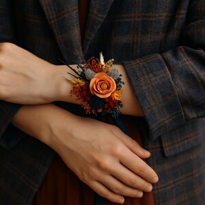Könnte beinhalten: Ein Handgelenk-Corsage mit einer orangefarbenen Rose und getrockneten Blumen in Rot, Gelb und Blau. Es ist mit einem marineblauen Band befestigt. Die Person trägt einen karierten Blazer. Ein floraler Schmuck.