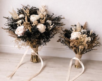 May include: Two bouquets of dried flowers. The larger bouquet features white roses, black foliage, and tan accents, tied with a burlap wrap and cream ribbon. The smaller bouquet has a similar design. Both are set against a white wall and light wood floor.
