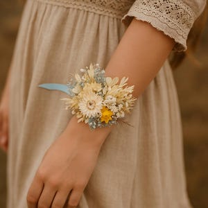 Könnte beinhalten: Ein Handgelenkscorsage aus getrockneten Blumen in Weiß-, Gelb- und Blautönen. Die Corsage ist mit einem hellblauen Band gebunden und enthält eine Vielzahl getrockneter Blumen, darunter Gänseblümchen, Schleierkraut und getrocknete Blätter.