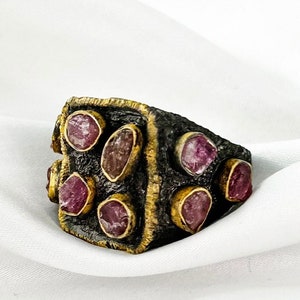 Op de afbeelding: Een massieve zilveren ring met een vierkant oppervlak, bezet met acht roze edelstenen. De ring heeft een getextureerde afwerking en een gouden rand.