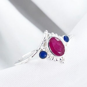 Może przedstawiać: Srebrny pierścionek z owalnym, rubinowym kamieniem pośrodku i dwoma mniejszymi, okrągłymi, niebieskimi kamieniami. Pierścionek ma ozdobny, ażurowy wzór.
