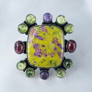 Puede incluir: Un anillo con una gran piedra preciosa verde y morada rodeada de piedras preciosas más pequeñas de color verde, morado y rojo.