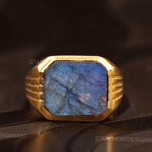 May include: A gold-toned ring with a rectangular, iridescent blue gemstone. The ring has a thick band with etched details. The gemstone displays a range of blue hues, creating a striking contrast against the gold.