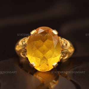 Peut inclure: Une bague en or avec une pierre précieuse jaune ovale. La bague présente un motif orné et filigrané sur le bandeau. La pierre précieuse est facettée et reflète la lumière. Le texte "STATEMENTDESIGNSART" est visible.