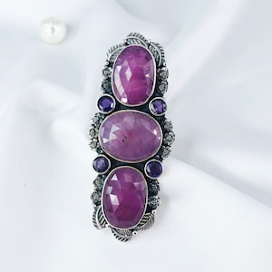 May include: A silver ring with three oval-shaped gemstones. The center stone is a large, pink gemstone, and the two smaller stones on either side are purple.