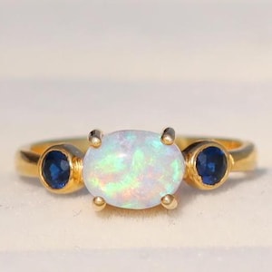 Könnte beinhalten: Ein goldener Ring mit einem weißen Opal-Edelstein und zwei kleineren blauen Saphiren auf jeder Seite.