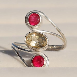 May include: A silver ring featuring three stones. The top stone is a round, red gemstone. The middle stone is an oval, yellow gemstone. The bottom stone is a round, red gemstone.