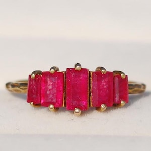 Puede incluir: Un anillo de oro con cinco piedras preciosas rectangulares de color rojo brillante. Las piedras están dispuestas en una fila en la parte superior del anillo. El anillo tiene una banda texturizada. Un diseño llamativo.