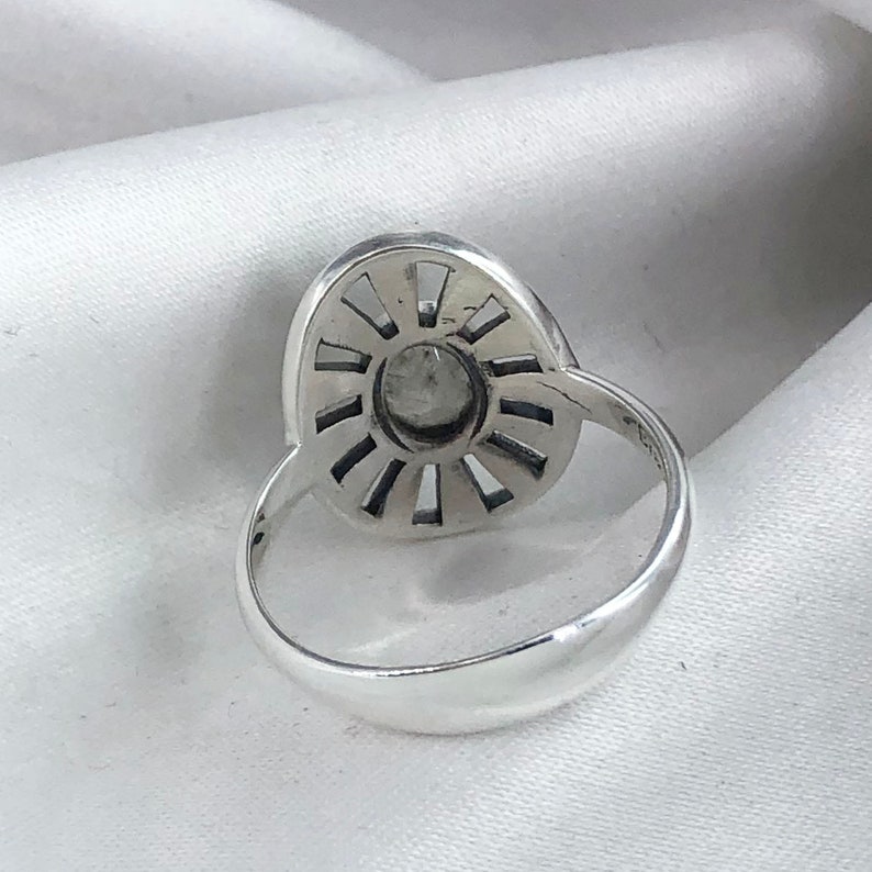 May include: A silver ring featuring a large, oval-shaped stone set within a sunburst design. The stone is encircled by a ring of smaller stones.