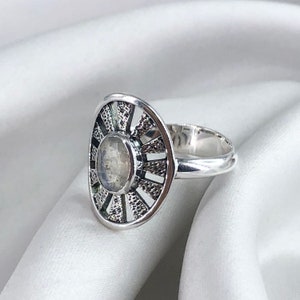 May include: A silver ring featuring a moonstone set within a circular design. The ring has a delicate, intricate pattern surrounding the stone.