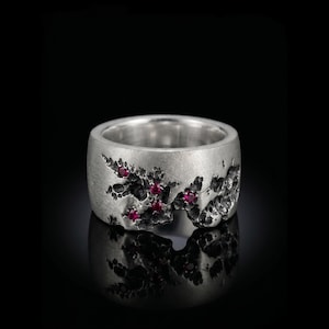 May include: A wide silver band ring featuring a textured, cratered surface with an organic, uneven edge. The ring is set with five small, round, vibrant red rubies embedded within the recessed, dark-toned crevices of the metal finish.