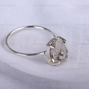 K&ouml;nnte beinhalten: Ein silberner Ring mit einem klaren, facettierten, rohen Diamanten. Der Diamant ist in einem schlichten, modernen Design gefasst.