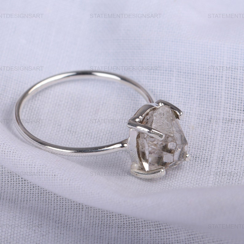 K&ouml;nnte beinhalten: Ein silberner Ring mit einem klaren, facettierten, rohen Diamanten. Der Diamant ist in einem schlichten, modernen Design gefasst.