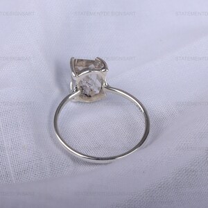 K&ouml;nnte beinhalten: Ein silberner Ring mit einem klaren, facettierten Edelstein. Der Ring liegt auf einem wei&szlig;en Stoffhintergrund.