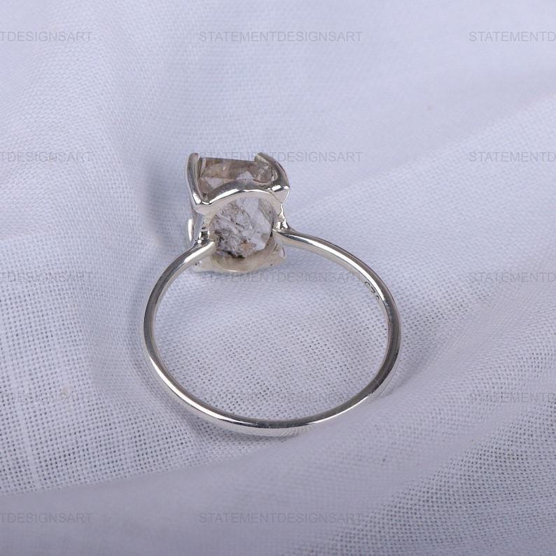 K&ouml;nnte beinhalten: Ein silberner Ring mit einem klaren, facettierten Edelstein. Der Ring liegt auf einem wei&szlig;en Stoffhintergrund.