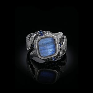 Könnte beinhalten: Ein Ring mit einem rechteckigen blauen Stein, der in ein dunkles, strukturiertes, oxidiertes Metallband gefasst ist. Die Fassung ist mit kleinen, runden, tiefblauen Steinen verziert, die in die raue, unebene Oberfläche des Metalls eingelassen sind.