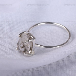 K&ouml;nnte beinhalten: Ein silberner Ring mit einem klaren, facettierten, rohen Diamanten. Der Diamant ist in einem schlichten, modernen Design gefasst.