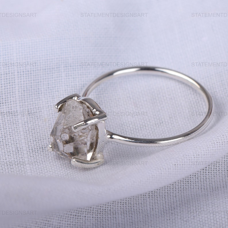 K&ouml;nnte beinhalten: Ein silberner Ring mit einem klaren, facettierten, rohen Diamanten. Der Diamant ist in einem schlichten, modernen Design gefasst.
