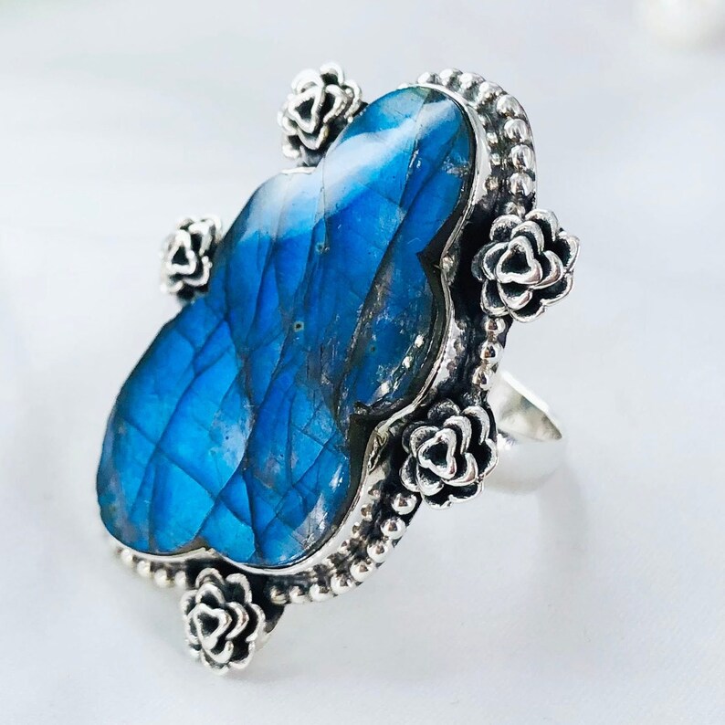 May include: A silver ring with a large blue labradorite gemstone set in a floral design. The ring is adorned with small silver flowers around the gemstone.