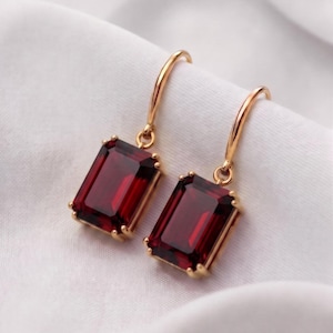 Può includere: Un paio di orecchini pendenti in oro con pietre preziose rosso scuro dal taglio rettangolare a smeraldo. Ogni pietra è incastonata in una montatura in oro a quattro griffe, sospesa a un gancio in oro ricurvo.