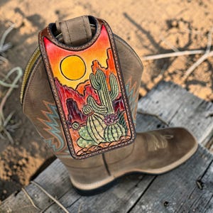 Könnte beinhalten: Eine braune Leder-Handyhülle mit einem Wüstenlandschaftsdesign mit Kakteen, einer untergehenden Sonne und Bergen. Die Hülle ist an einem braunen Cowboy-Stiefel befestigt, der auf einer Holzoberfläche ruht.