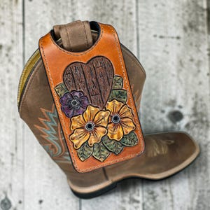 Könnte beinhalten: Ein handgefertigter Leder-Stiefelriemen in Braun- und Orangetönen, verziert mit einem geschnitzten Holzherz und floralem Design. Der Riemen hat zwei gelb-orangefarbene Blumen mit schwarzen Zentren und eine lila Blume. Der Riemen liegt auf einem braunen Cowboy-Stiefel.