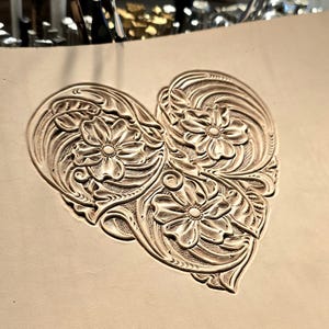 Peut inclure: Gros plan sur une sculpture en cuir en forme de cœur. Le motif présente des motifs floraux détaillés avec des feuilles et des pétales tourbillonnants. La sculpture est de couleur beige clair naturel, mettant en valeur les détails en relief.