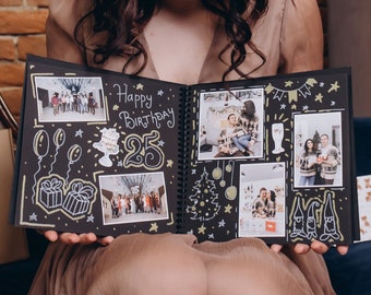 May include: A woman is holding a black photo album open to a page with the words "Happy Birthday 25" written on it. The album is filled with photos and drawings.