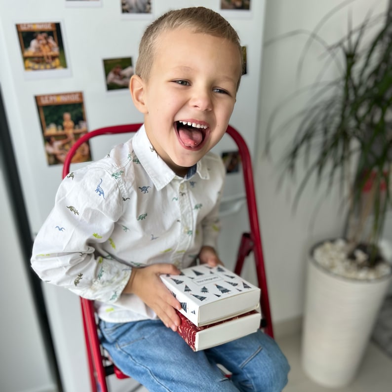May include: A young boy wearing a white button-down shirt with a dinosaur print sits on a red chair and holds two boxes wrapped in white paper with a black and white print of Christmas trees. The boxes are stacked on top of each other.
