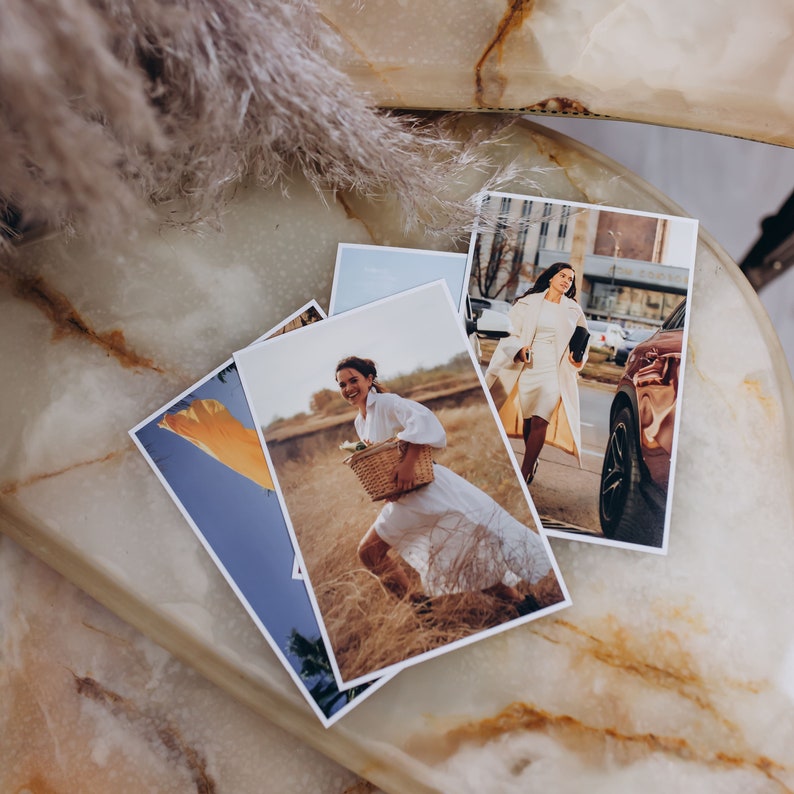 May include: Three photographs are displayed on a marble surface. The first photo shows a woman in a white dress holding a basket in a field. The second photo shows a woman in a white dress and a beige coat walking down a city street. The third photo shows a car parked on a city street.