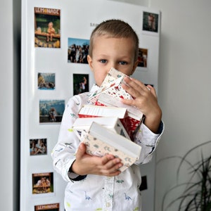 May include: A young boy holds a stack of colorful, patterned boxes in front of his face. The boxes are stacked in a pyramid shape and have a variety of designs, including flowers, geometric patterns, and stripes.