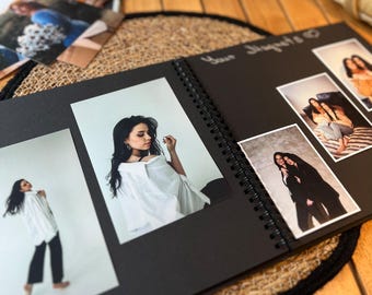 Puede incluir: Un álbum de fotos negro con encuadernación en espiral. El álbum está abierto en una página con varias fotos de personas. El texto "Your Journey" está escrito en la parte superior de la página.