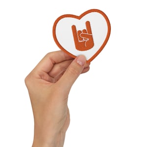 Könnte beinhalten: Ein weißer herzförmiger Patch mit einer orangefarbenen gestickten Rock'n'Roll-Handgeste. Der Patch wird in einer Hand gehalten.